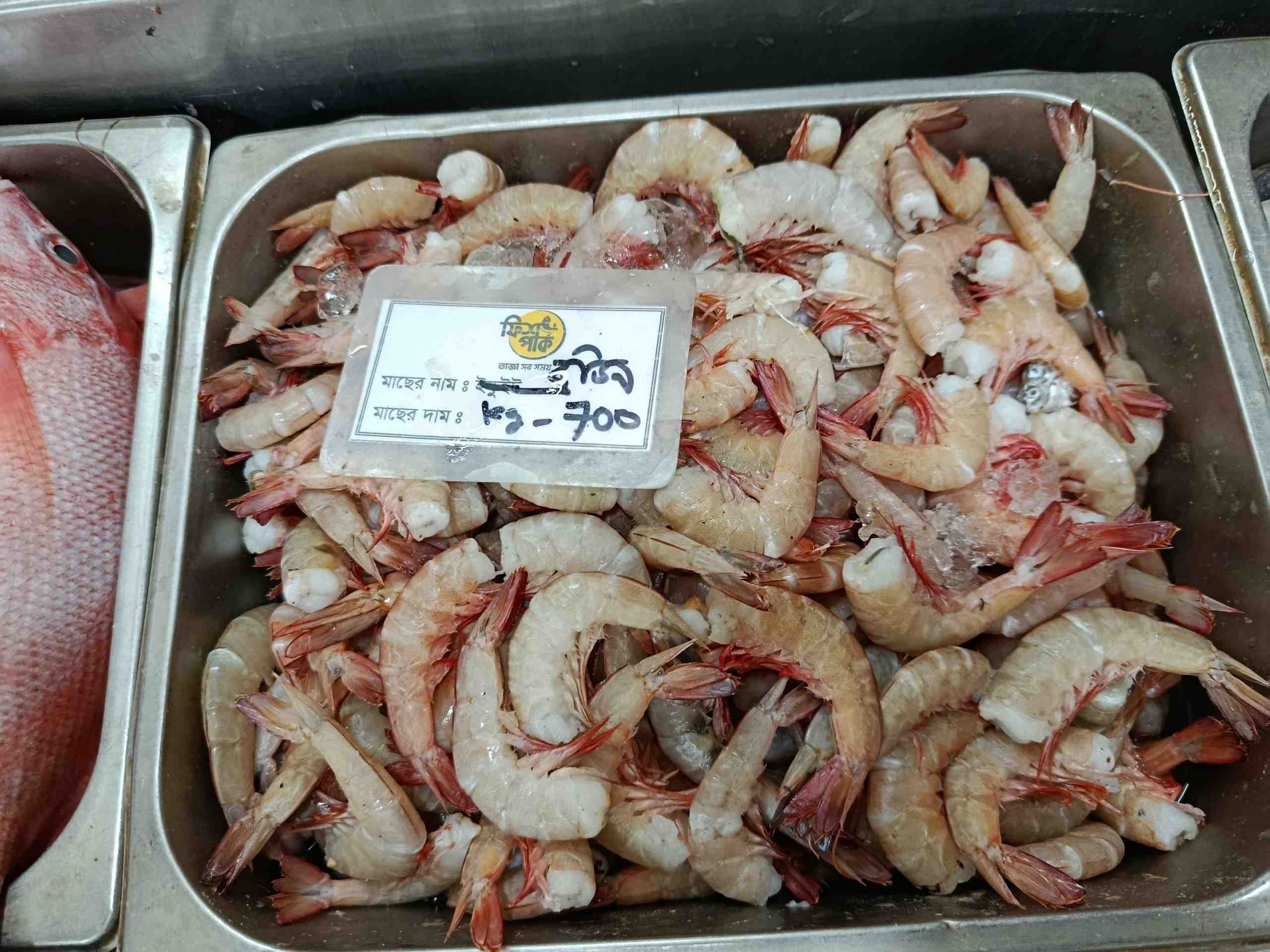 Fresh headless brown shrimp with reddish tails displayed with ice in a metal market tray at Fish Park