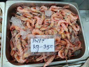 Fresh whole Redfin shrimp with bright pinkish-red shells displayed with ice and a price tag in a metal market tray