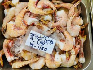 A top-down view of a metal bin filled with fresh, raw small white shrimp mixed with crushed ice, featuring a hand-written price tag on top.