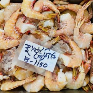 A top-down view of a metal bin filled with fresh, raw small white shrimp mixed with crushed ice, featuring a hand-written price tag on top.